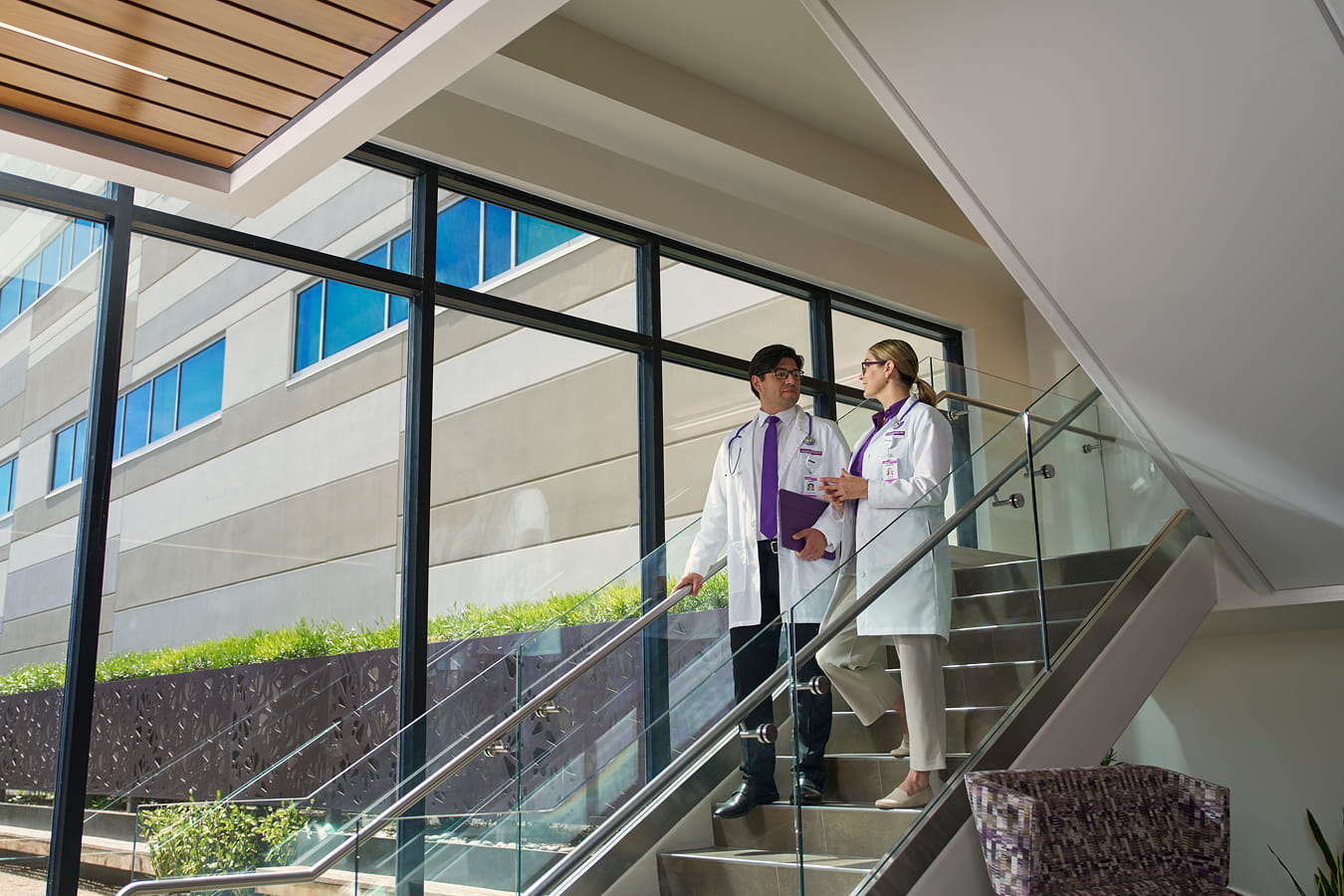 Two HonorHealth physicians in white coats converse while walking down a staircase inside a modern, sunlit healthcare facility.