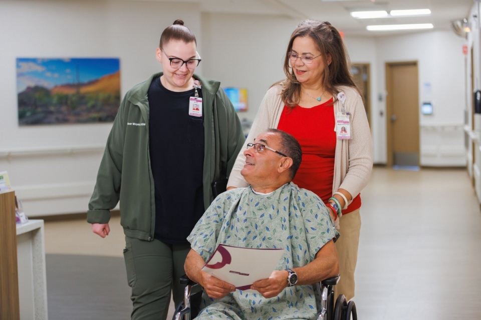 Two HonorHealth team members walk with a patient in a wheelchair down a hospital hallway, smiling and providing support after surgery.
