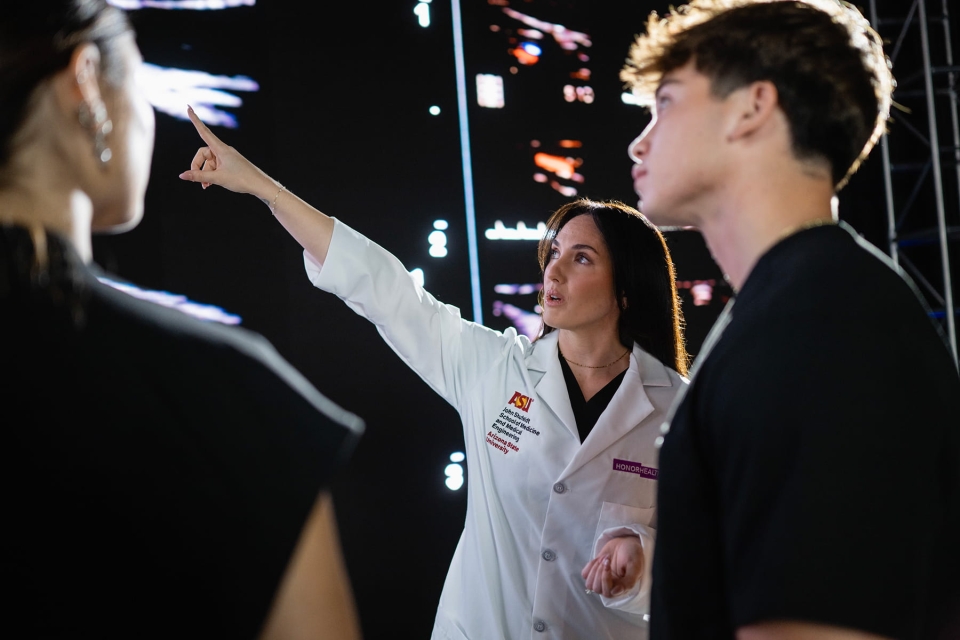 A medical educator in a white coat with an HonorHealth and ASU School of Medicine embroidery points toward a large digital screen while speaking with two students.