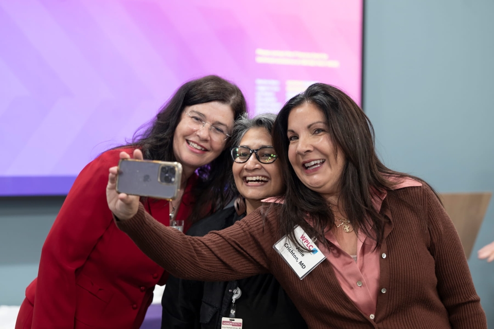 Three members of the Women Physician Leadership Council smile and take a selfie together with a smartphone at an event.