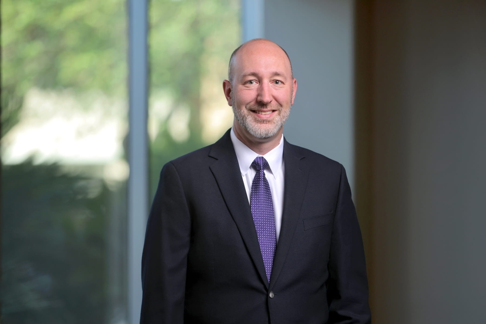Professional headshot of Dr. Brian Sidoti smiling, wearing a dark suit jacket, white dress shirt, and purple pattern tie against a blurred office background.
