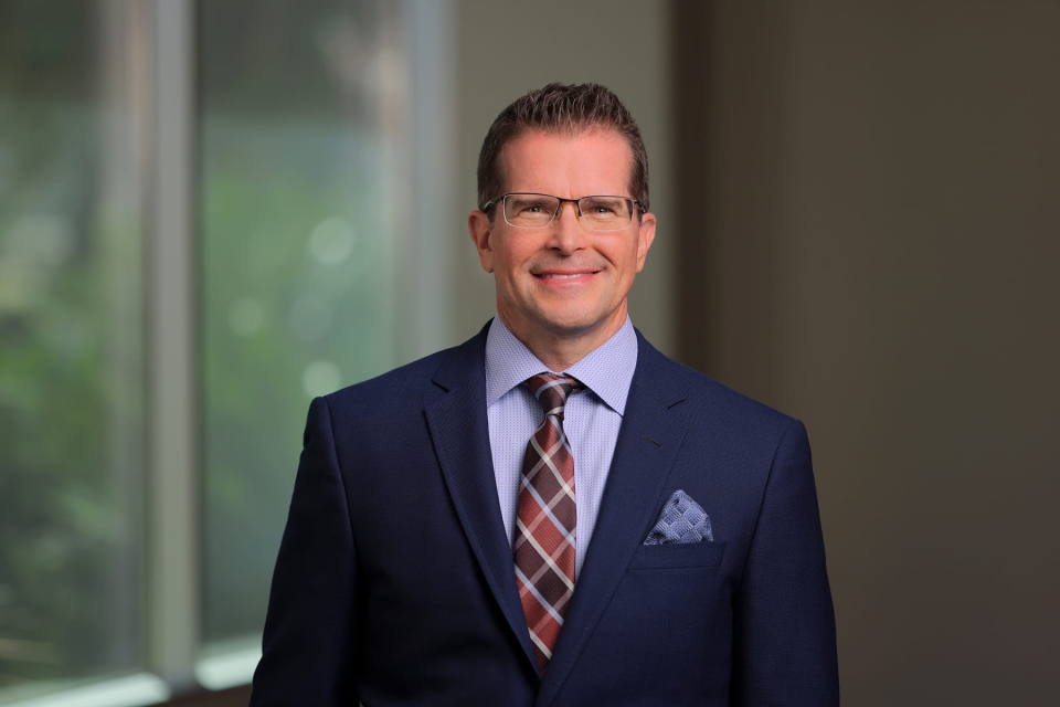 Professional headshot of Dr. John Pope smiling, wearing glasses, a dark blue suit jacket, light patterned shirt, plaid tie and pocket square against a blurred background.