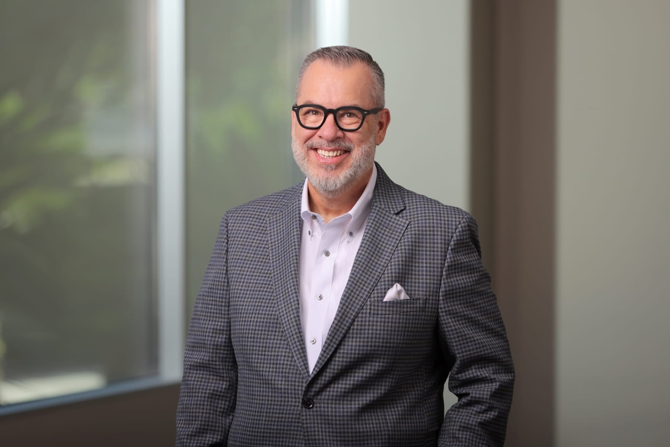 Professional headshot of Dr. Kevan Pickrel wearing black-framed glasses and a patterned grey suit jacket with a pocket square, smiling against a soft office background.