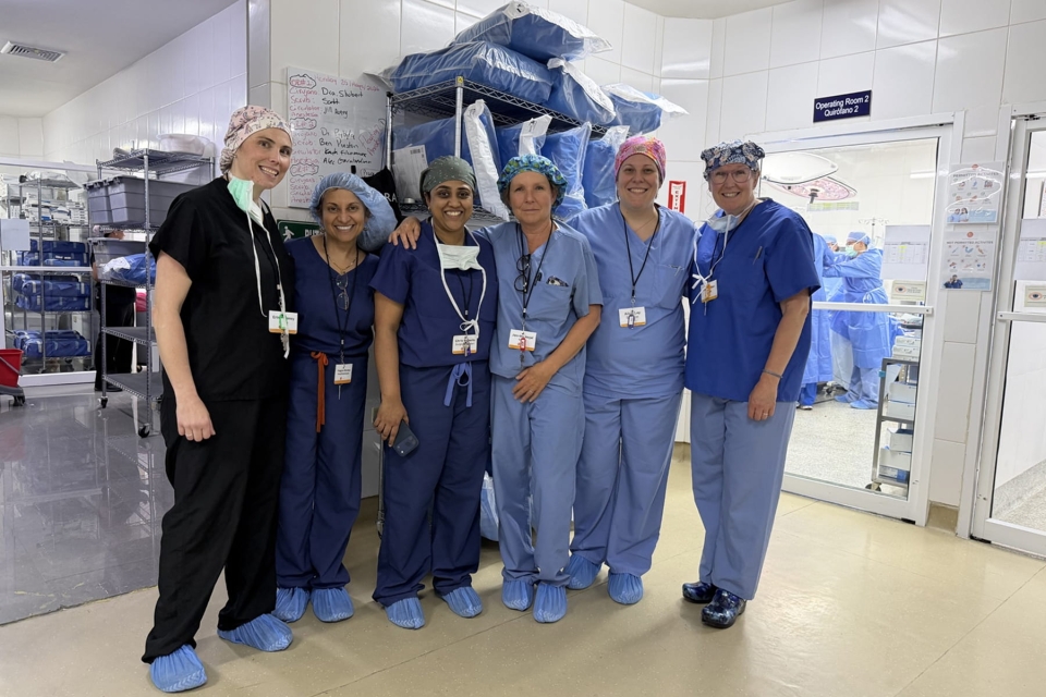 Six medical professionals wearing scrubs and surgical caps pose together in a hospital hallway outside an operating room during a medical mission in Honduras.