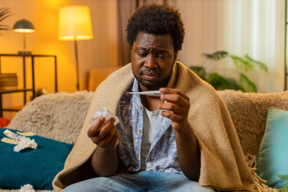HonorHealth Primary Care - Man takes temperature with thermometer while sitting on couch with a blanket draped over his shoulders.