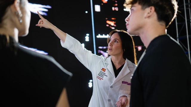 A medical educator in a white coat with an HonorHealth and ASU School of Medicine embroidery points toward a large digital screen while speaking with two students.