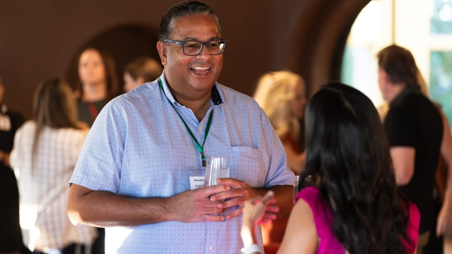 A smiling male physician in glasses holds a glass and chats with a woman physician at a Medical Staff social.