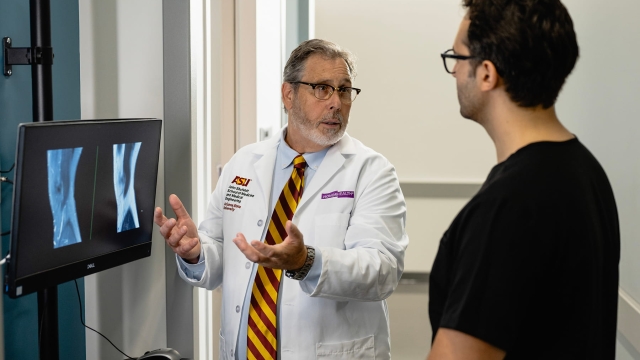 A doctor wearing an ASU and HonorHealth embroidered white coat speaks with a colleague while gesturing toward medical imaging displayed on a monitor.
