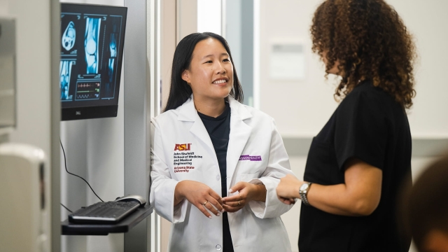 A smiling physician in a white lab coat co-branded with Arizona State University and HonorHealth logos speaks with a colleague near a wall-mounted monitor displaying medical scans.