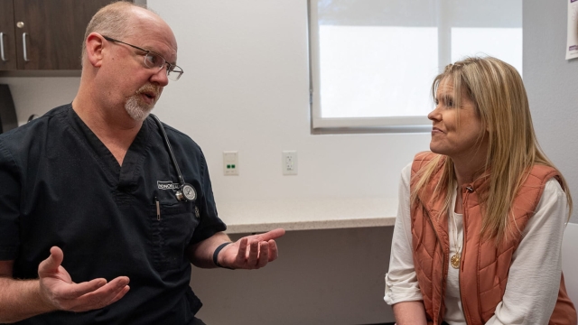 Dr. Swain wearing dark scrubs and a stethoscope gestures with his hands while speaking with a female patient in an exam room.