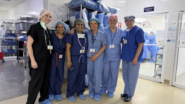 Six medical professionals wearing scrubs and surgical caps pose together in a hospital hallway outside an operating room during a medical mission in Honduras.