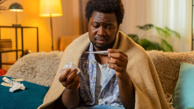 HonorHealth Primary Care - Man takes temperature with thermometer while sitting on couch with a blanket draped over his shoulders.