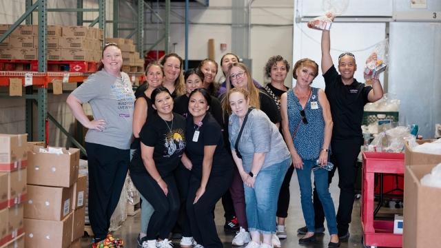 A large group of HonorHealth Desert Mission volunteers smile and pose together inside a warehouse surrounded by food donation boxes.