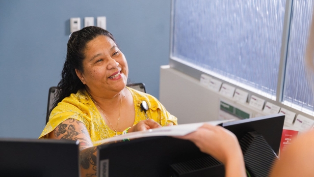 A smiling team member in a yellow top sitting behind a reception counter and handing paperwork over a computer monitor to a visitor.
