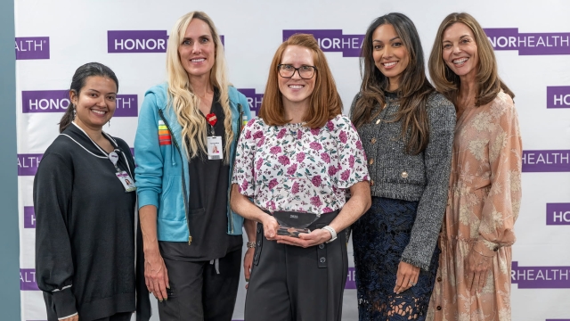 Five smiling women pose for a group photo against a white backdrop featuring purple HonorHealth logos. The Trailblazer Award recipient in the center, wearing glasses and a floral blouse, holds a clear trophy.